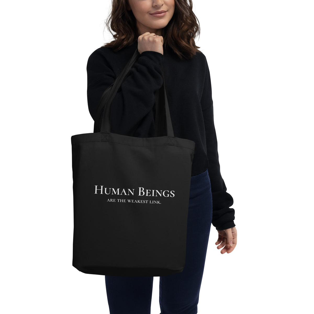 Model is standing against a white background holding front-facing graphic eco tote bag in color black. Featuring a bold statement, 'Human Beings Are The Weakest link', in bold white print, from What Was Said (Statement Apparel + More). 