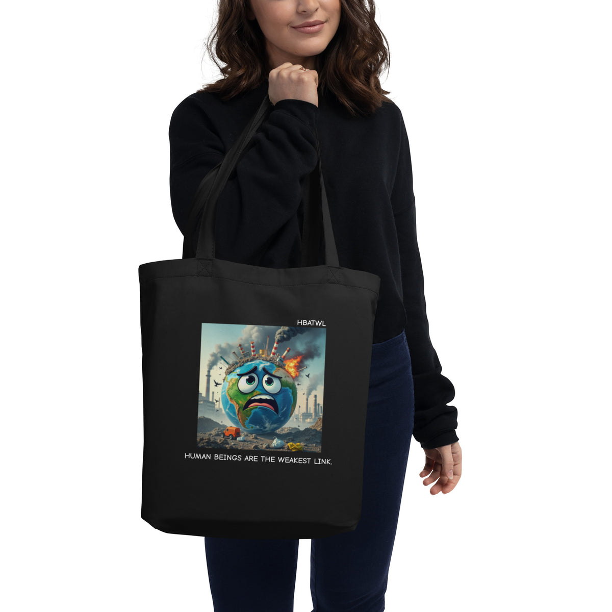 Model is standing against white background holding front-facing graphic eco tote bag in color black, featuring an animated earth with facial expression of concern over being negatively effected by human pollution. Bold statement on the bag reads, 'Human Beings Are The Weakest link.', in white print. From What Was Said (Statement Apparel + More). 