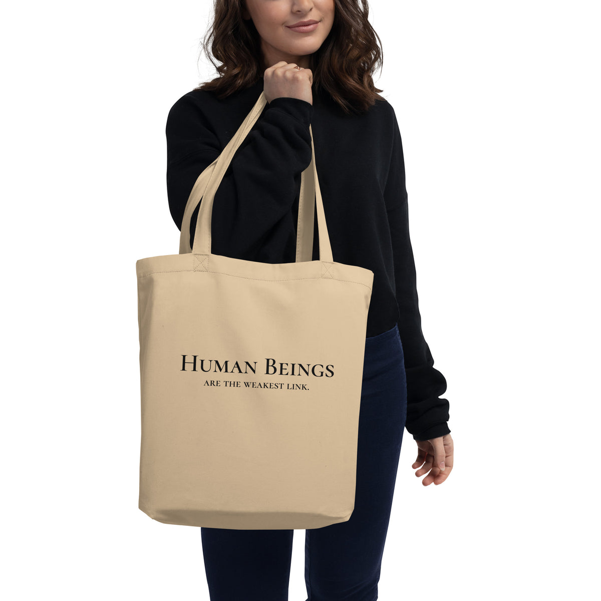 Model is standing against a white background holding front-facing graphic eco tote bag in color oyster beige. Featuring a bold statement, 'Human Beings Are The Weakest link', in bold black print, from What Was Said (Statement Apparel + More). 