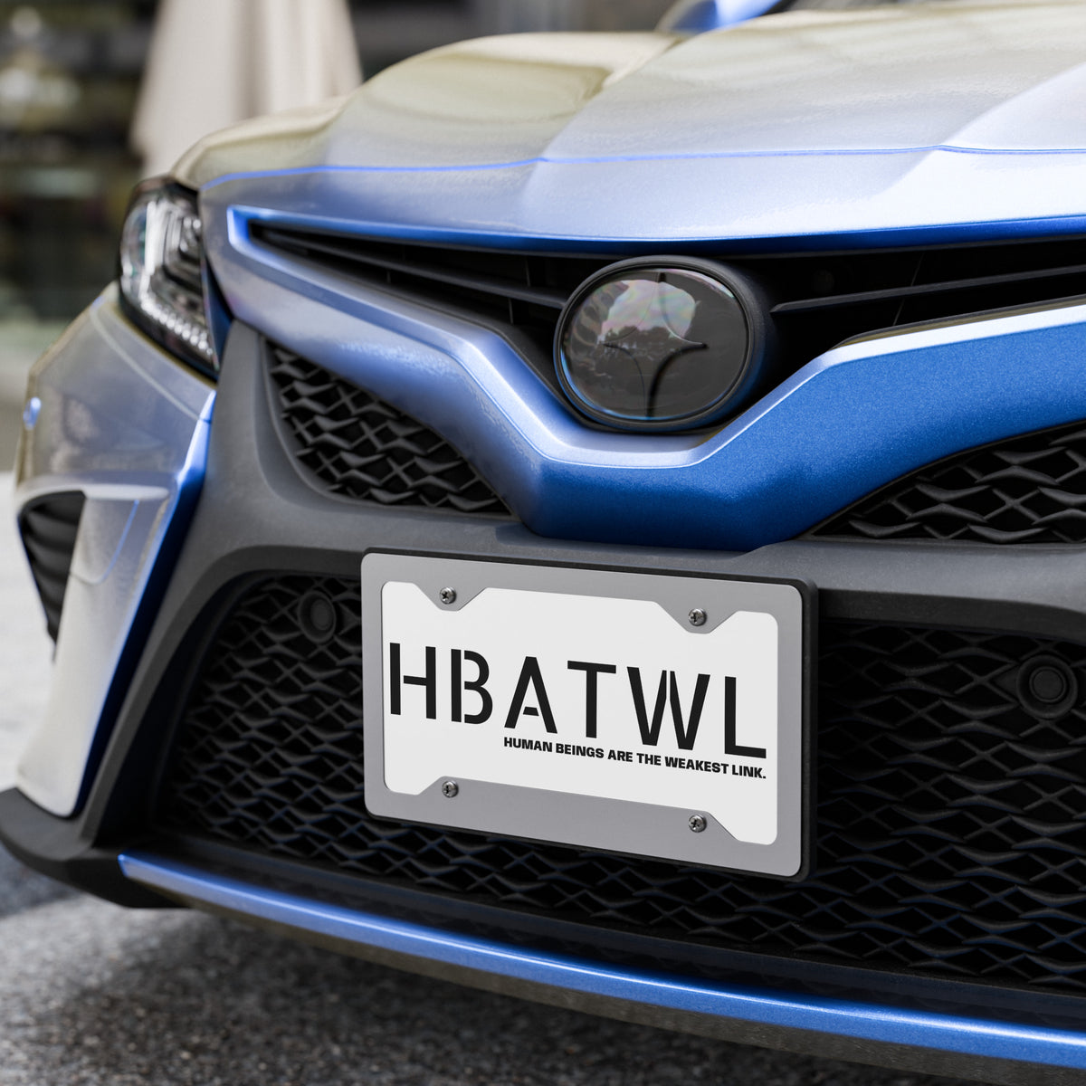 Front facing graphic vanity plate in color white, featuring the initialism 'HBATWL' and the statement 'HUMAN BEINGS ARE THE WEAKEST LINK.' in bold black print, affixed to a car's front grill. From What Was Said (Statement Apparel + More). 