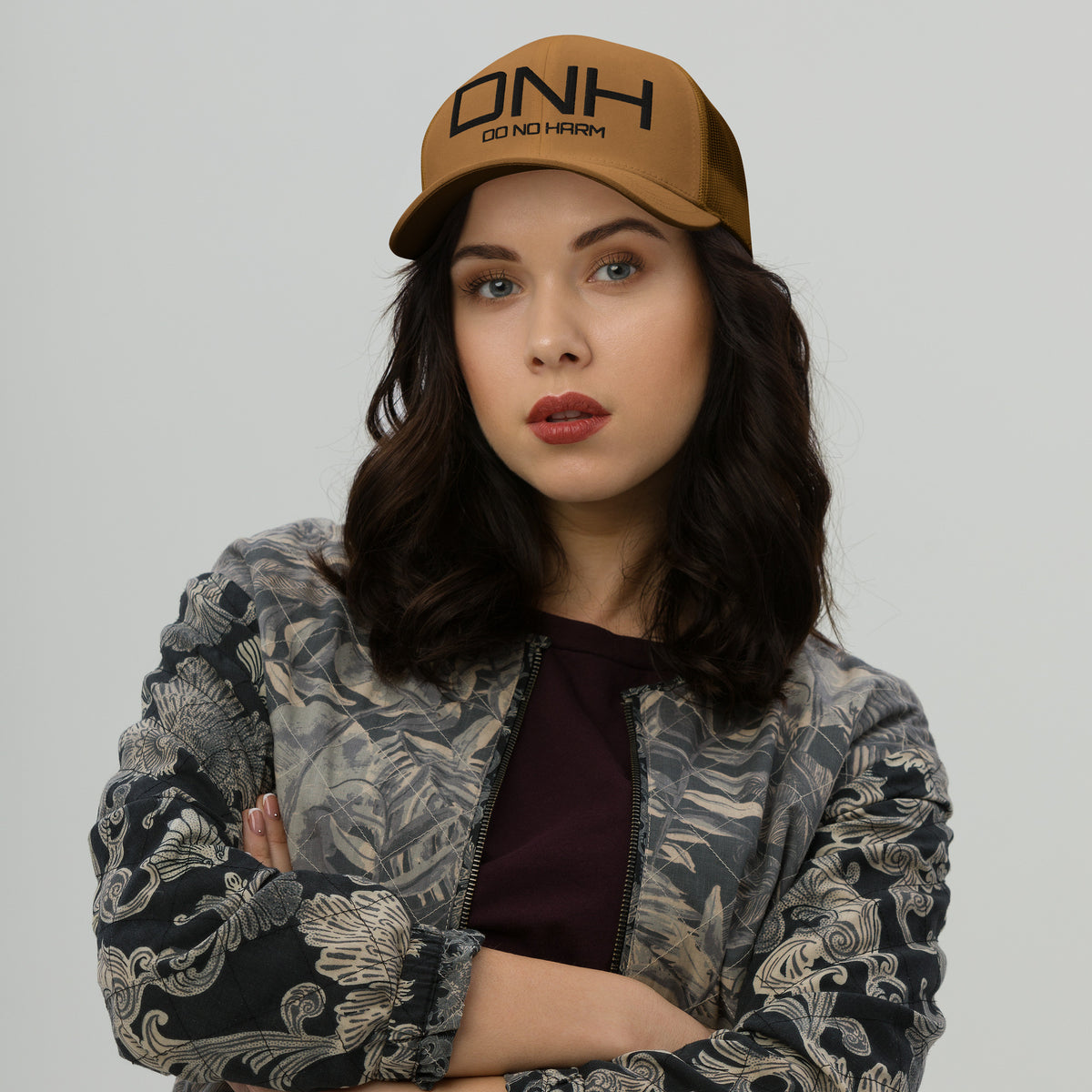 Model is wearing front facing graphic trucker hat in color caramel, featuring the initialism 'DNH', and the statement 'DO NO HARM,' in bold black embroidery. From What Was Said (Statement Apparel + More).