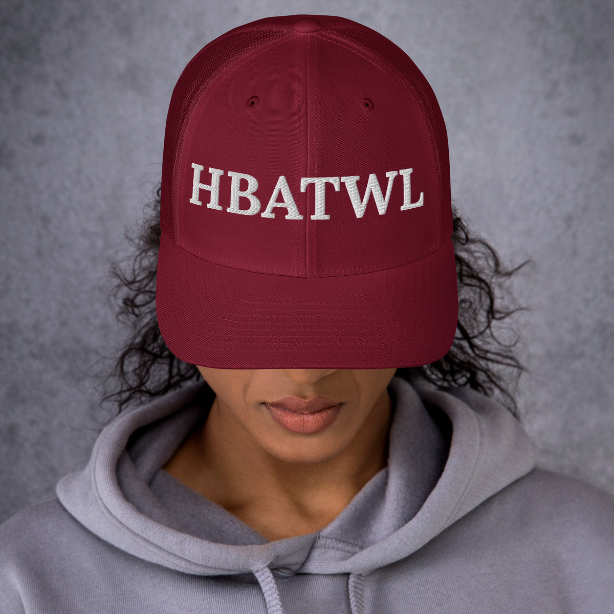 Model is wearing front facing graphic trucker hat in color cranberry red, featuring the initialism 'HBATWL', from What Was Said (Statement Apparel + More).