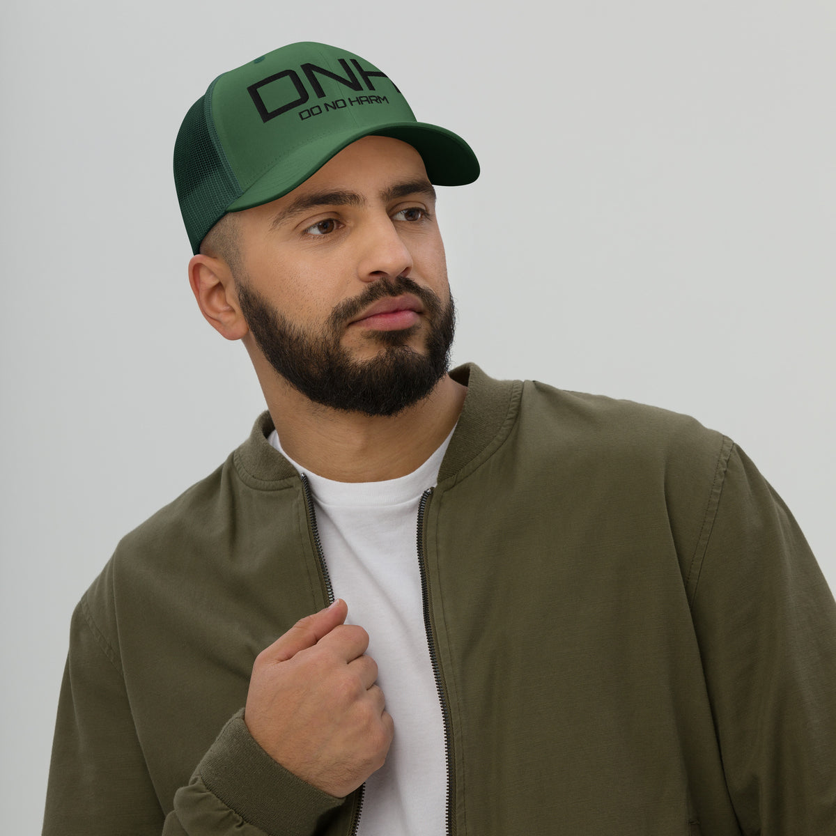 Model is wearing front facing graphic trucker hat in color evergreen, featuring the initialism 'DNH', and the statement 'DO NO HARM,' in bold black embroidery. From What Was Said (Statement Apparel + More).