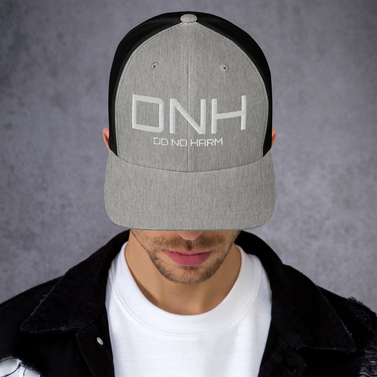 Model is wearing front facing graphic trucker hat in color heather grey and black, featuring the initialism 'DNH', and the statement 'DO NO HARM,' in bold white embroidery. From What Was Said (Statement Apparel + More).