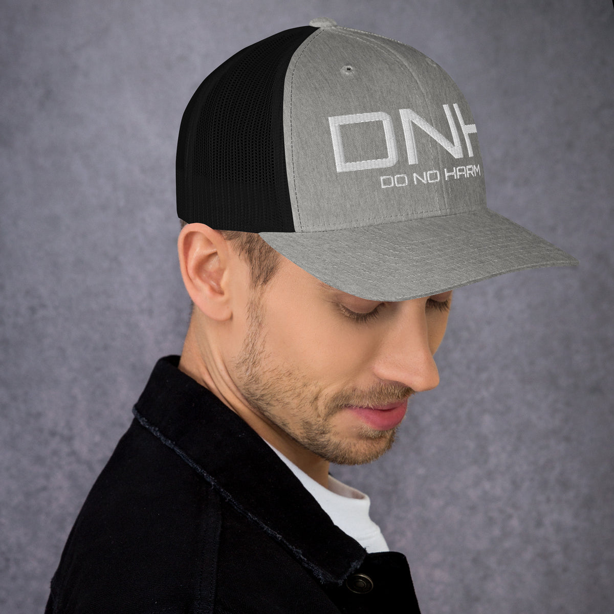 Model is wearing right side front facing graphic trucker hat in color heather grey and black, featuring the initialism 'DNH', and the statement 'DO NO HARM,' in bold white embroidery. From What Was Said (Statement Apparel + More).
