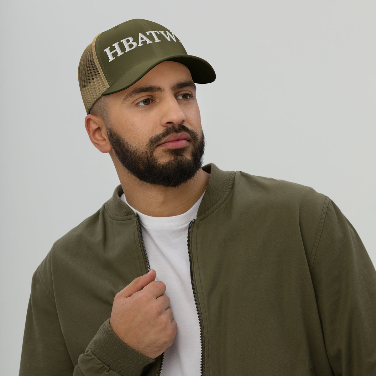 Model is wearing front facing graphic trucker hat in color moss and khaki, featuring the initialism 'HBATWL', from What Was Said (Statement Apparel + More).