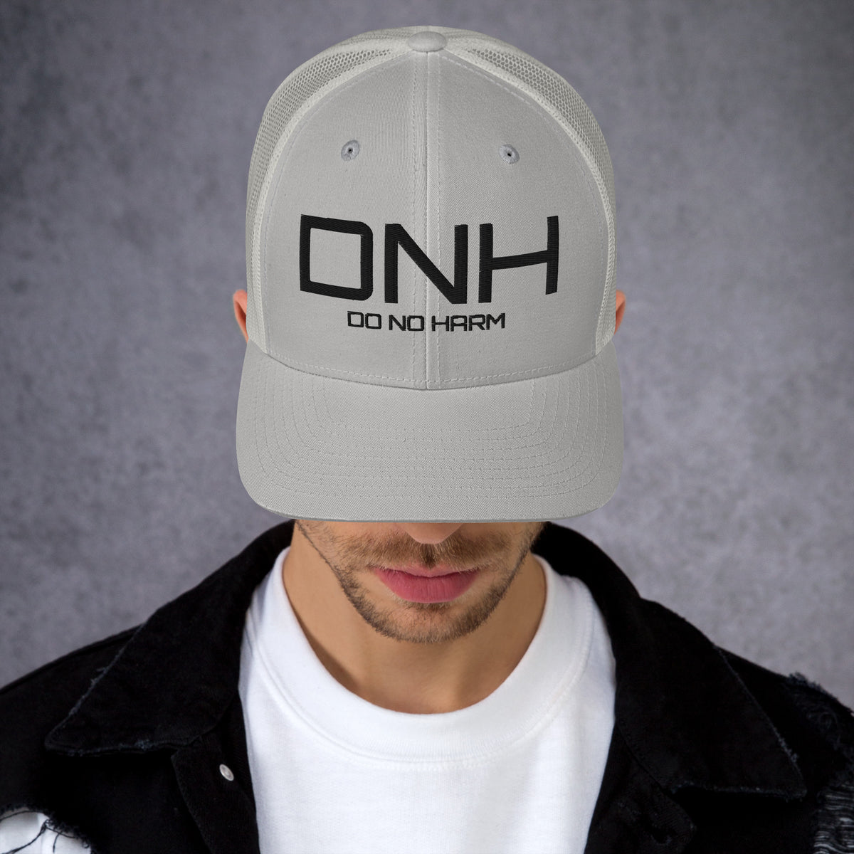 Model is wearing front facing graphic trucker hat in color silver light grey, featuring the initialism 'DNH', and the statement 'DO NO HARM,' in bold black embroidery. From What Was Said (Statement Apparel + More).