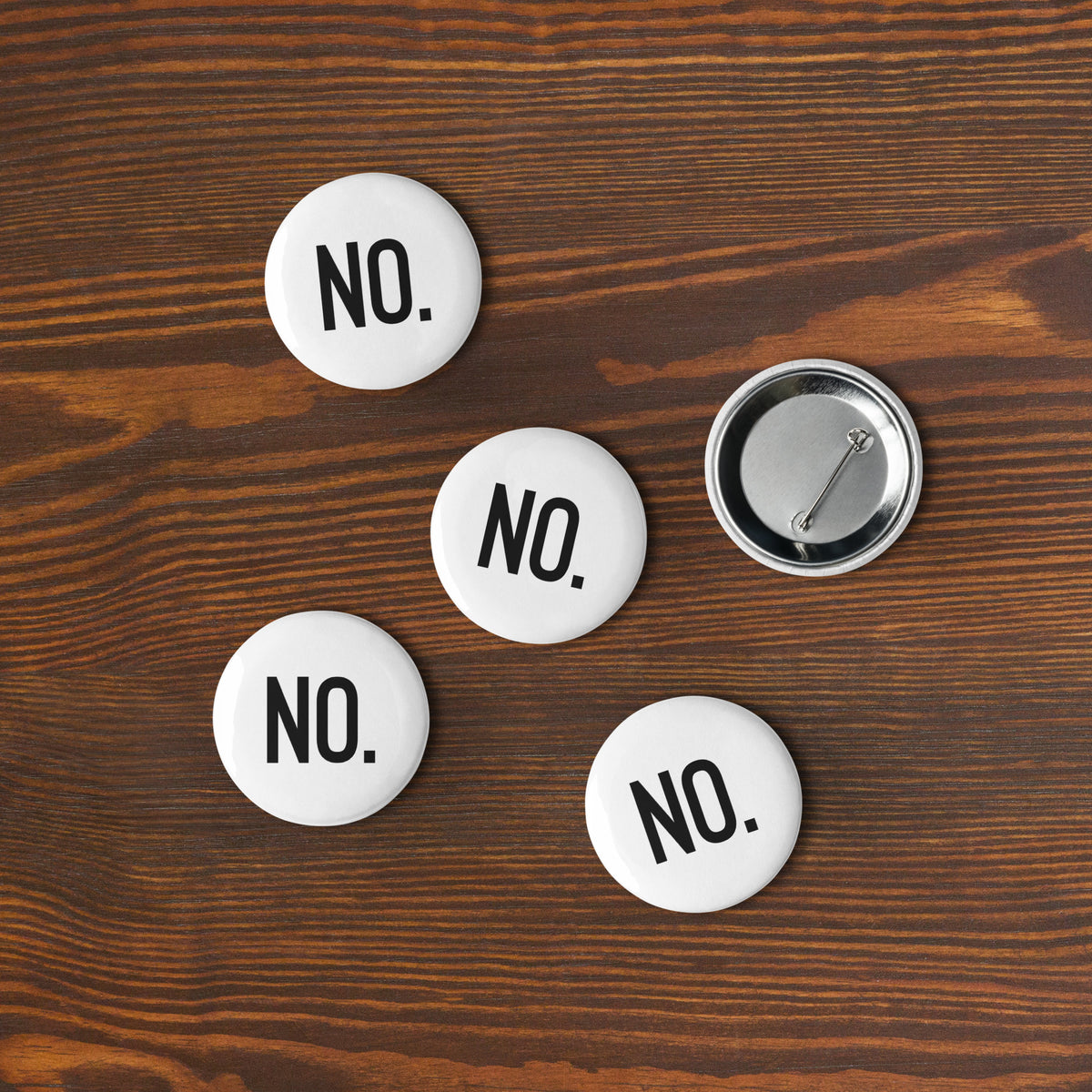 Front and back facing graphic pin buttons against a woodgrain desk background. Buttons are in color white and size 2.25 inches. Featuring safety pin type clasp, and the statement NO in bold black print. From What Was Said (Statement Apparel + More). 