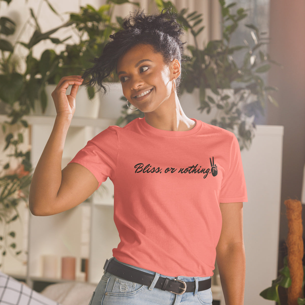 Model is wearing front facing graphic t-shirt in color coral silk, featuring statement, 'Bliss, or nothing,' in bold black cursive print. With a hand showing the peace sign at the end of the statement. From What Was Said (Statement Apparel + More).