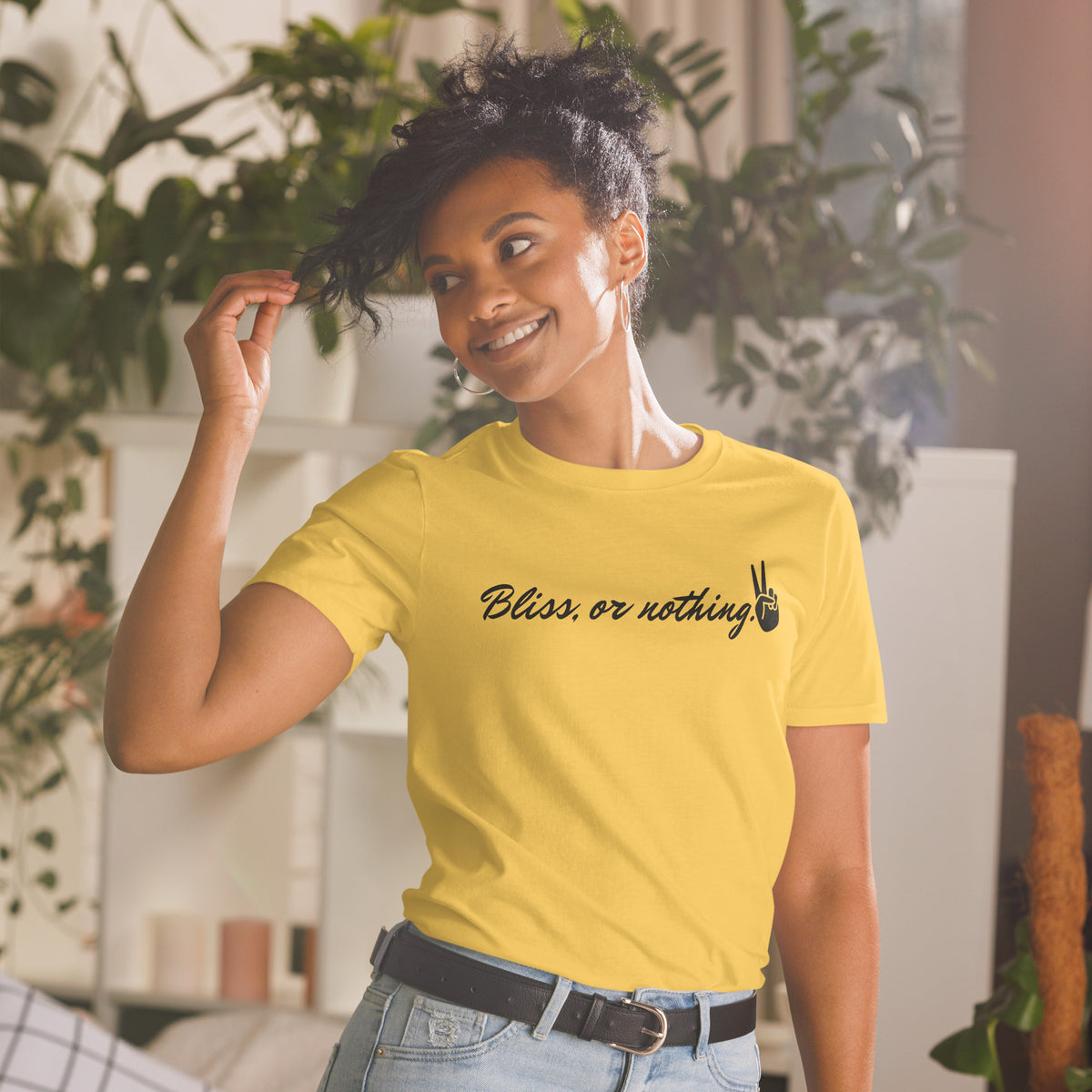 Model is wearing front facing graphic t-shirt in color daisy yellow, featuring statement, 'Bliss, or nothing,' in bold black cursive print. With a hand showing the peace sign at the end of the statement. From What Was Said (Statement Apparel + More).