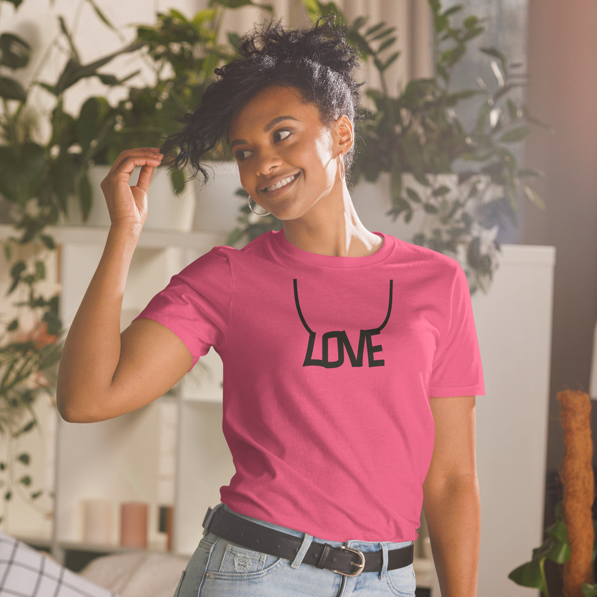 Model is wearing front facing graphic T-shirt in color heliconia pink, featuring printed image of the written word 'LOVE' as a necklace, in black print. From What Was Said (Statement Apparel + More).