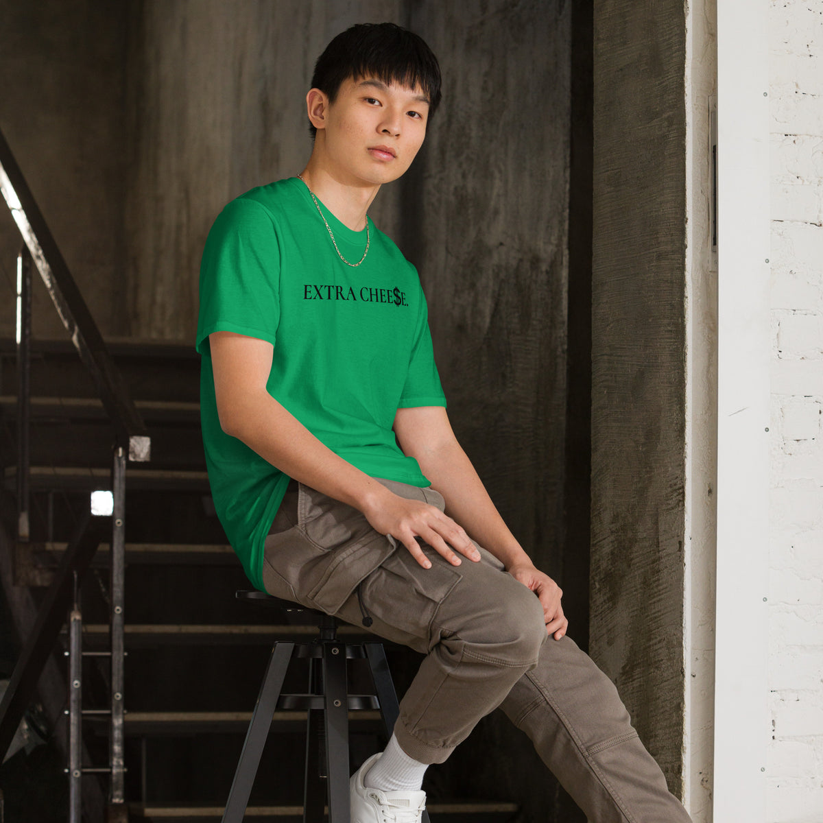 Model is wearing right front facing graphic T-shirt in color irish-green, featuring statement 'EXTRA CHEESE', with dollar sign symbol in place of the letter S in the word cheese, in black print. From What Was Said (Statement Apparel + More).