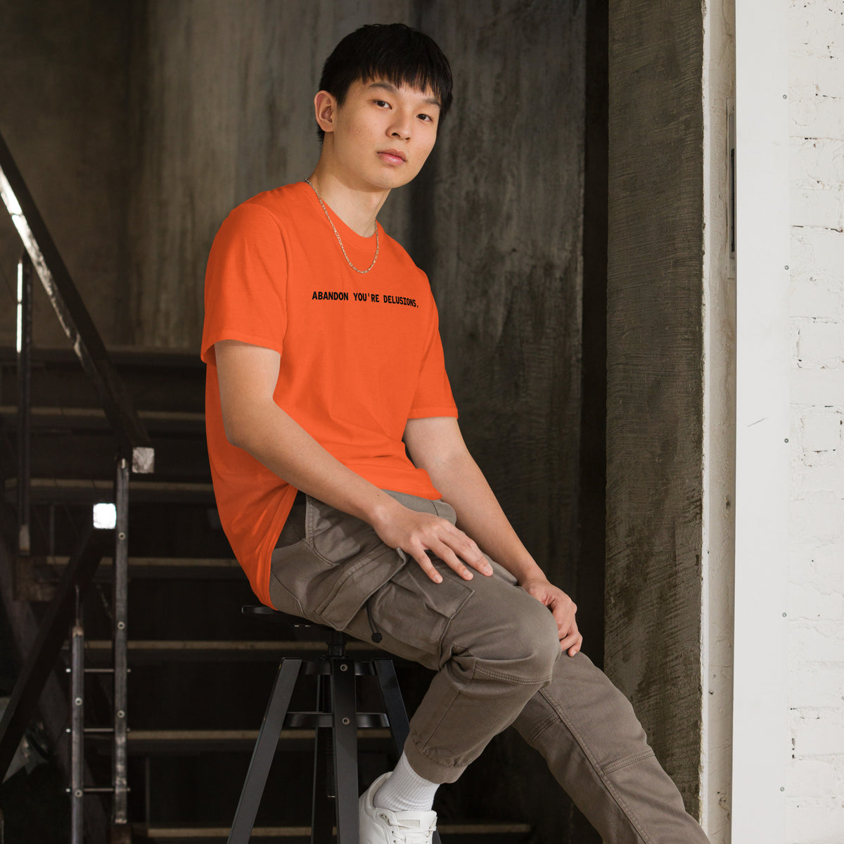 Model wearing right front facing graphic t-shirt in color orange, featuring statement, 'ABANDON YOU'RE DELUSIONS', in bold black print. From What Was Said (Statement Apparel + More).