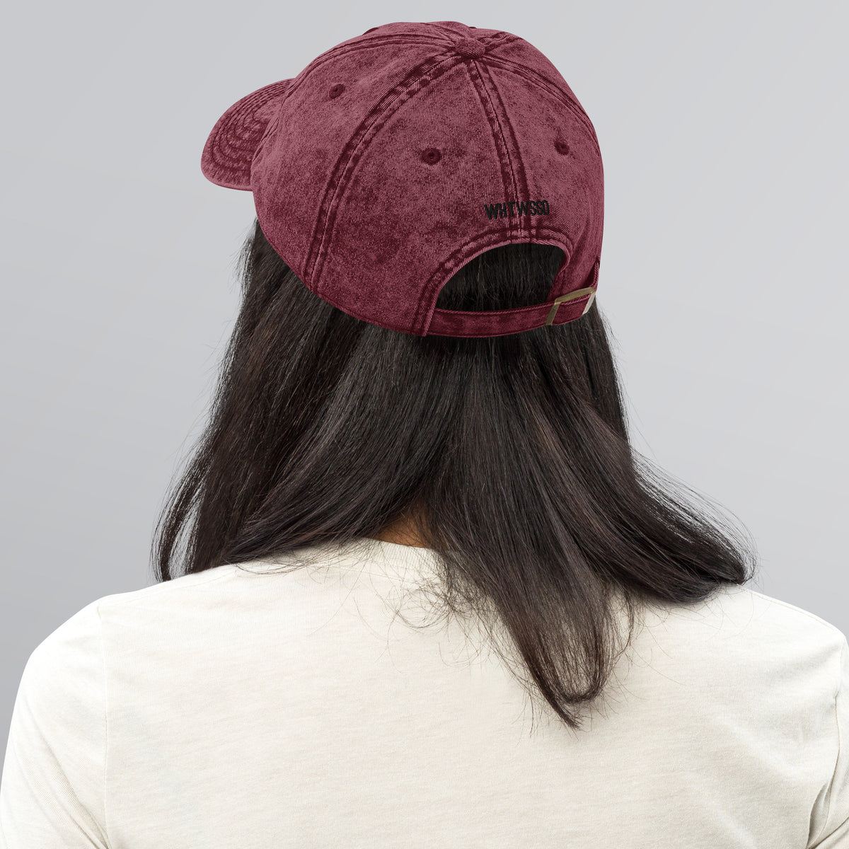 Model is wearing back facing vintage graphic cotton twill cap in color charcoal maroon red wash, featuring view of hats metal snap buckle closure and brand acronym WHTWSSD in black print. From What Was Said (Statement Apparel + More).